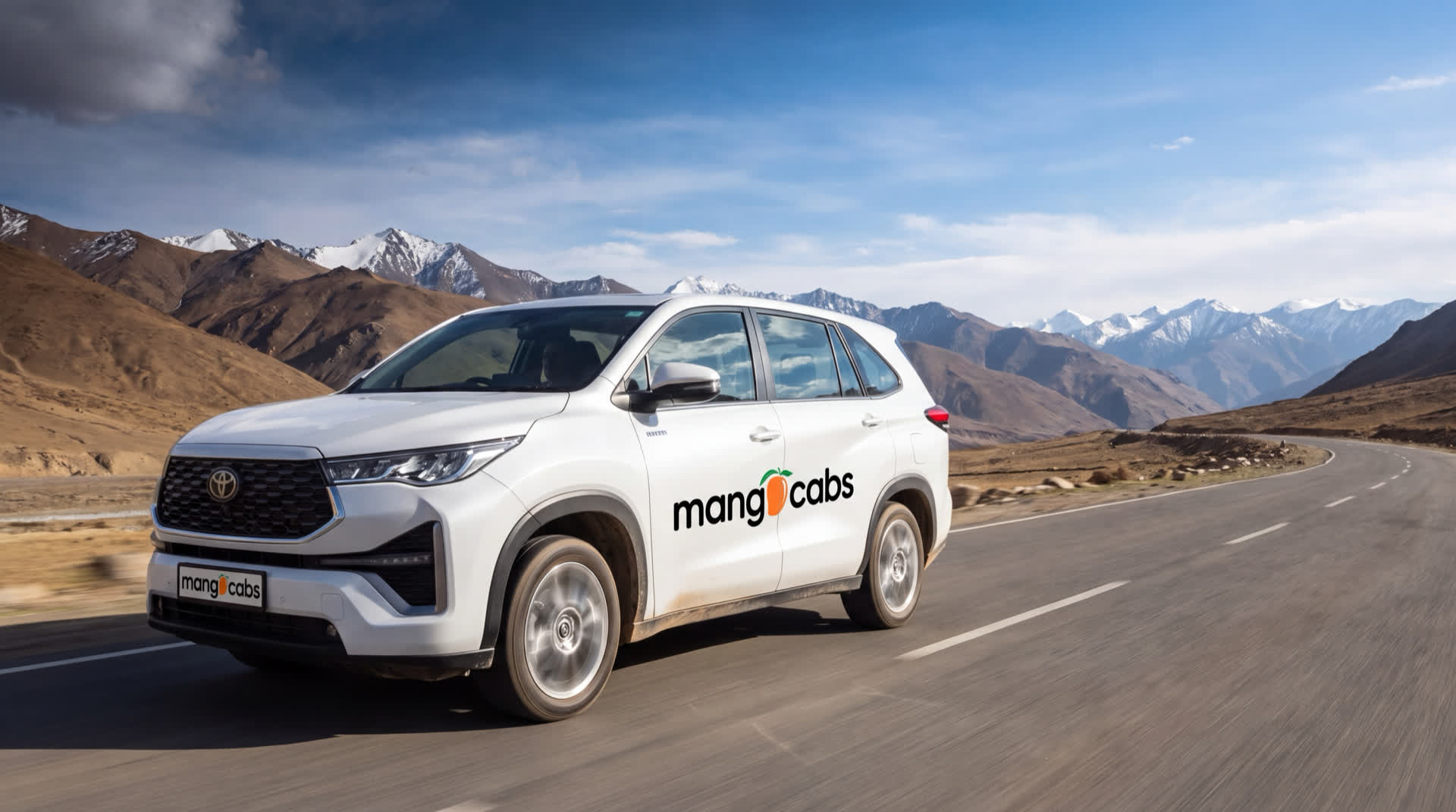 A white Mango Cabs Toyota Innova Hycross on an open Ladakh mountain highway, snow-capped Himalayan peaks and bare high-altitude ridges behind it